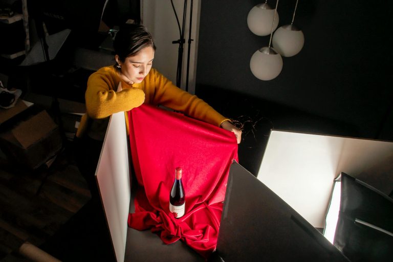 Photographer arranging a studio scene with a wine bottle and red fabric using controlled lighting.