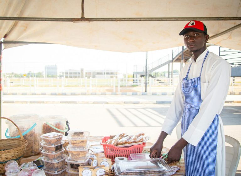 A young male vendor in Kaduna, Nigeria, selling baked goods at an outdoor market stall.